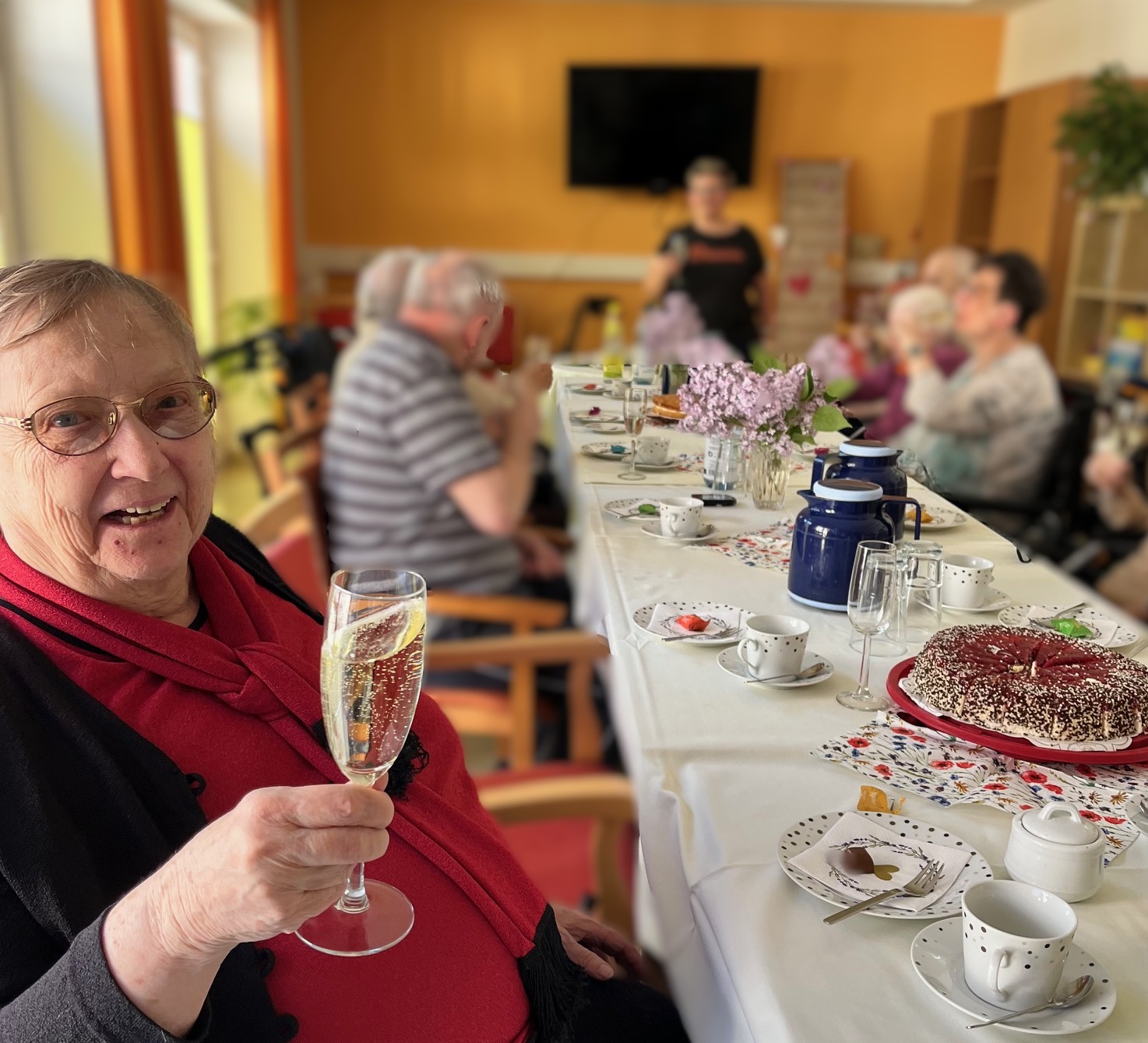 Senior*innen sitzen am Kaffeetisch, eine Seniorin ist im Vordergrund mit Sekt